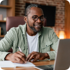 A middle-aged man smiles as he writes notes taken from his laptop.   